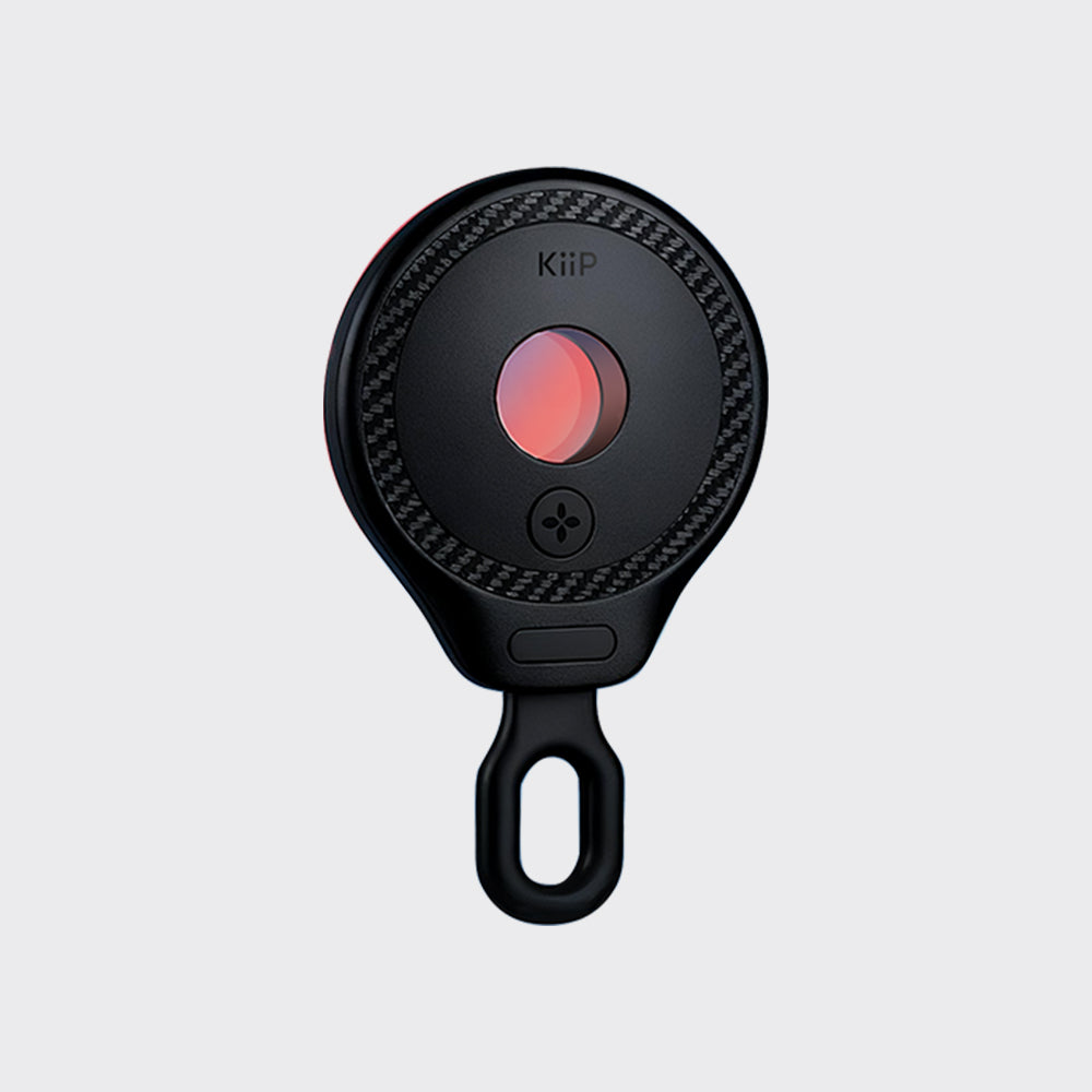 A black circular KiiP personal alarm with a carbon-fiber-textured outer ring, a glowing pink-red center button, and a hanging loop at the bottom, shown on a light gray background.